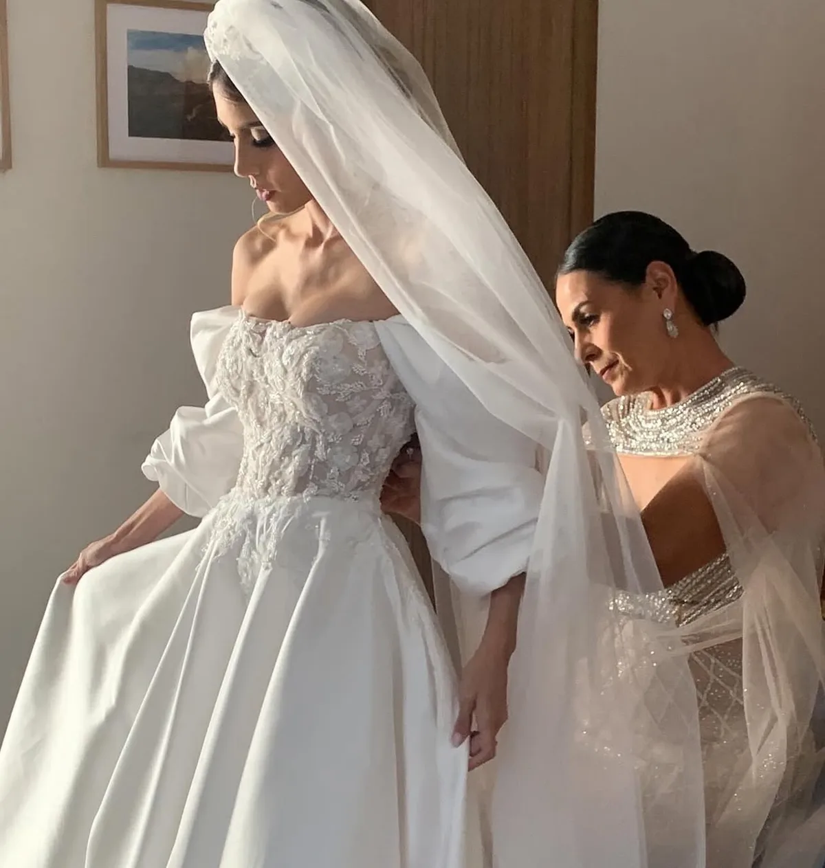 Novia de perfil con velo mientras le abrochan el vestido de pedrería